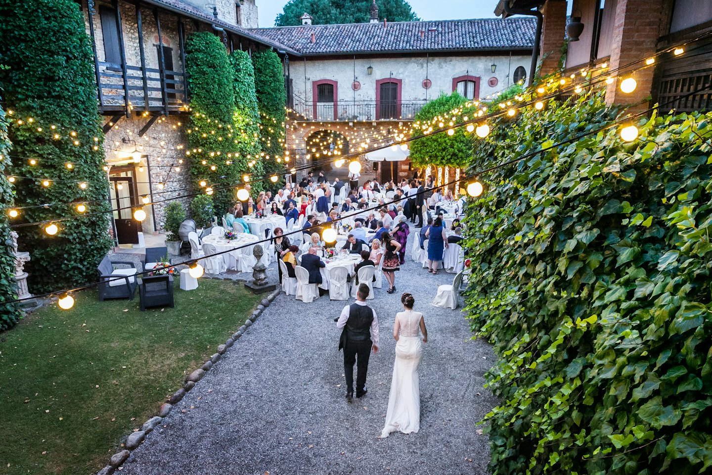 Newlyweds walk through a courtyard reception with globe lights.